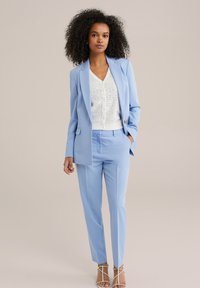 Ljust blå skräddarsydd kostym med en-bladblazer, slim-fit byxor och vit spetsblus. Sandaler med remmar i nude färg fulländar looken.
