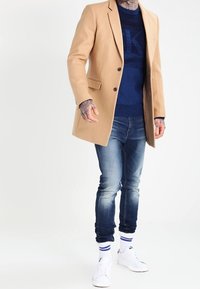 Kamelfärgad skräddarsydd kappa, mörkblå tröja, blekta blå jeans, vita sneakers, med blå och vita randiga strumpor. Detaljer inkluderar knappar.