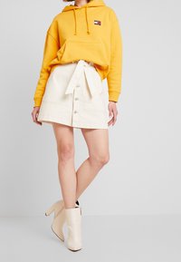 Sudadera amarilla con capucha y bolsillo frontal, combinada con una falda de mezclilla blanca que presenta un lazo delantero y cierre de botones, y botas de tacón blancas hasta el tobillo.