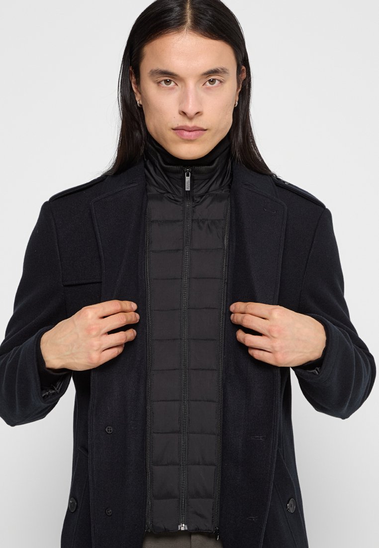 Cappotto di lana nero con chiusura a bottone e giacca interna imbottita con zip. Caratteristiche notevoli includono texture doppie e colletto alto.