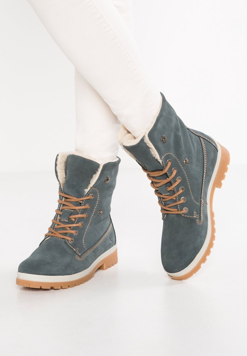 Bottes cheville en daim gris avec laçage à l'avant, accents couleur beige, doublure en fausse fourrure, et semelles en caoutchouc avec un motif de crampons robuste.