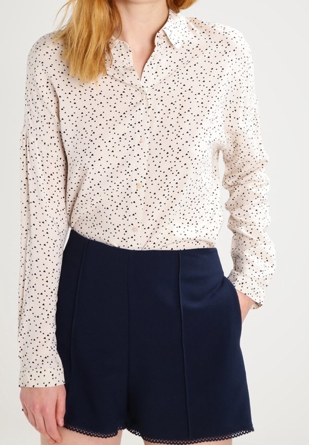 Femme portant un chemisier blanc à manches longues avec des pois noirs et un short bleu marine taille haute avec un ourlet festonné.