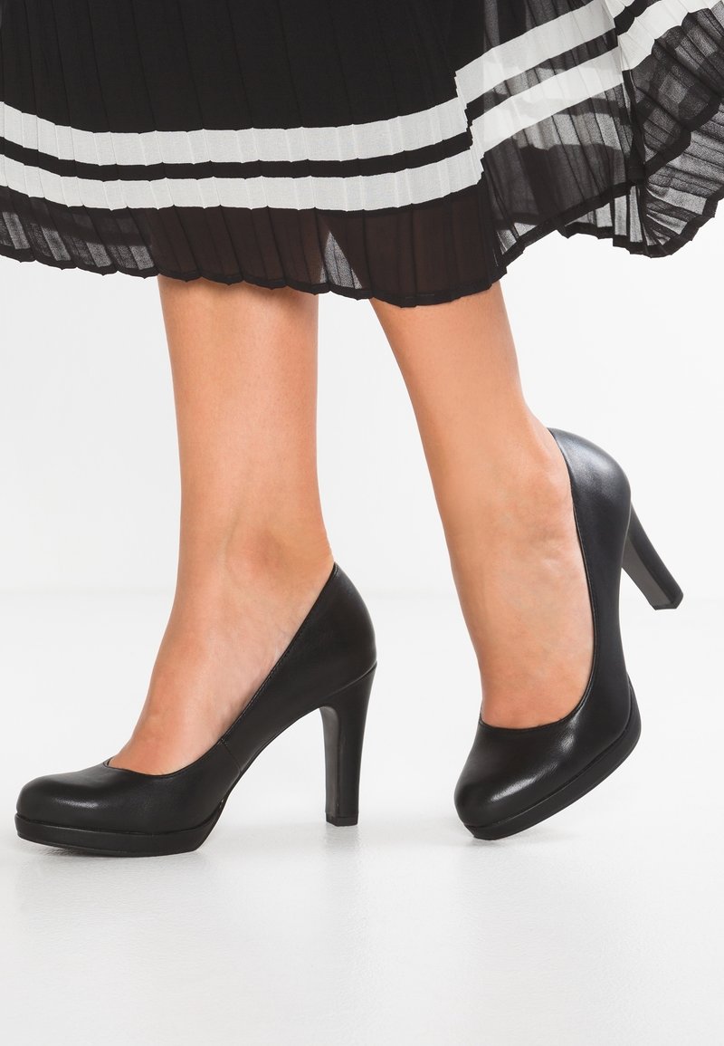 Jambes portant des talons hauts en cuir noir avec une jupe plissée noire présentant des rayures horizontales blanches.