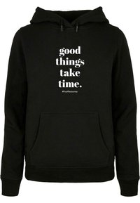 Haut à capuche noir en tissu doux, doté d'une poche kangourou. Le texte à l'avant indique "les bonnes choses prennent du temps" et inclut un hashtag.