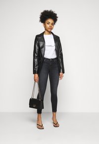 Black faux leather blazer over a white ruffled blouse, paired with dark skinny jeans and black handbag with chain strap. Model wears flip-flops.