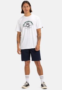 Camiseta blanca de algodón con estampado gráfico verde y texto, combinada con pantalones cortos azul marino. El modelo lleva zapatillas negras y calcetines blancos a rayas.