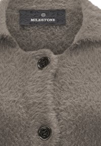 Nahaufnahme eines weichen grauen, flauschigen Mantels mit schwarzen Knöpfen und einem schwarzen Etikett der Marke Milestone im Kragen.
