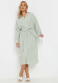 Lång, ljusgrön och vitrandig skjortklänning med knäppning framtill och bälte i midjan. Har långa ärmar och mjuk tygtextur. Bärs med vita sandaler.