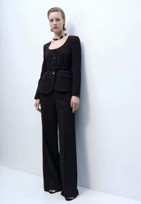 Blazer nero con scollatura rotonda, chiusura a bottone e cintura. Pantaloni neri larghi. Design semplice con accenti minimalisti.