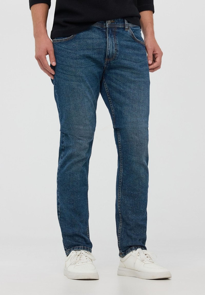 Niebieskie dżinsy o dopasowanym kroju, z pięcioma kieszeniami, z zamkiem błyskawicznym i subtelnym przetarciem. Połączone z białymi trampkami.