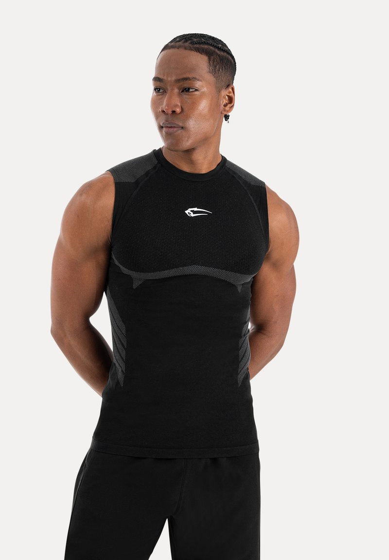 Homme athlétique avec des cheveux tressés portant un haut de compression noir sans manches ajusté avec un petit logo blanc, debout les mains derrière le dos.
