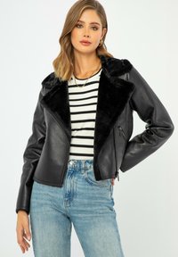 Giubbotto in ecopelle nera con un colletto morbido nero, tasche con cerniera e design aderente. Indossato sopra una camicia a righe e jeans blu.