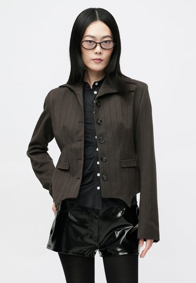 Barna csíkos szabott zakó, struktúrált gallérral, elöl gombokkal és két hajtókás zsebbel, fényes fekete sorttal párosítva.