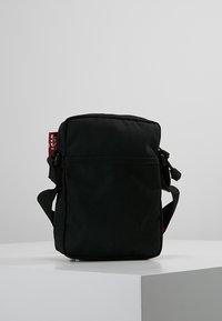Borsa a tracolla rettangolare nera con tracolla regolabile, tasca frontale con zip e tessuto testurizzato. Presenta un'etichetta con logo rossa sul lato.