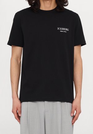 Person trägt ein schwarzes T-Shirt mit weißem Text "ICEBERG since 1974" auf der Brust, kombiniert mit hellgrauer Hose.