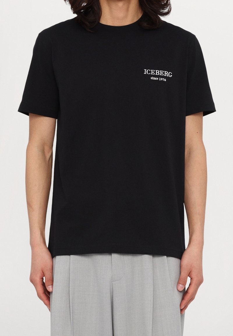 Personne portant un t-shirt noir avec le texte blanc "ICEBERG since 1974" sur la poitrine, associé à un pantalon gris clair.