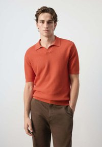 Jeune homme debout, une main dans la poche, portant un polo orange texturé à manches courtes et un pantalon marron sur un fond uni.
