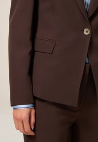 Blazer marrón a medida con un botón plateado, un bolsillo y una textura suave. Se puede ver un puño azul claro con rayas oscuras debajo.