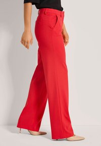 Rode, wijde broek met plooien, gemaakt van een gladde stof. Voorzien van zijzakken en een aansluitende tailleband. Gepaard met beige puntige hakken.