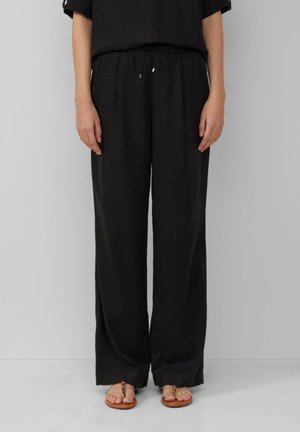 Nainen, jolla on päällään löysät mustat narupantsarit ja ruskeat sandaalit, joissa on ketjuyksityiskohta, seisomassa yksinkertaisen vaaleanharmaan taustan edessä.