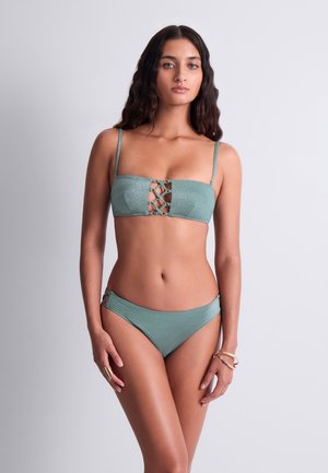 Femme aux longs cheveux foncés portant un bikini bandeau vert avec un détail lacé sur le haut et un bas assorti, debout devant un fond clair uni.