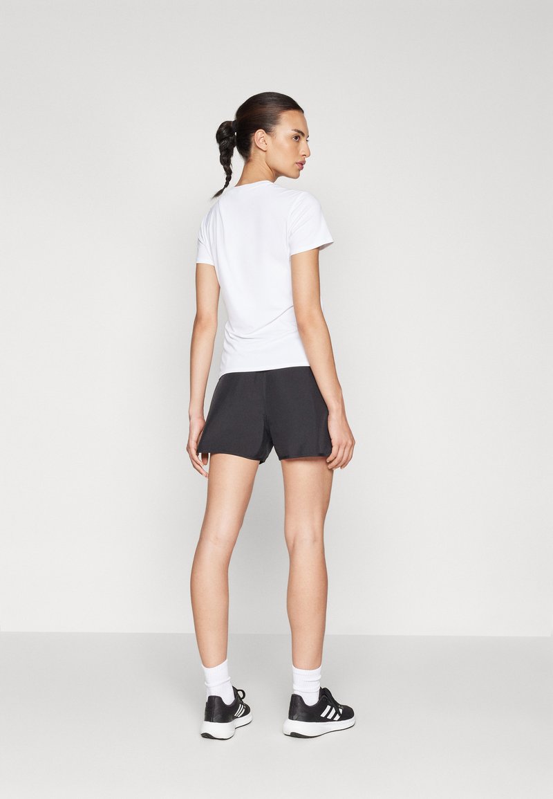 Camiseta blanca de manga corta combinada con shorts deportivos negros, vista desde la parte posterior. Se pueden ver zapatillas deportivas negras y calcetines blancos.