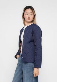 Junge Frau, die eine navyblaue Steppjacke über einem weißen Oberteil und blauen Jeans trägt, mit einer Hand in der Tasche steht und zur Seite schaut.