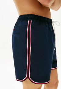 Bañadores de marinero con cintura elástica, cordón blanco y rayas laterales en rojo, blanco y azul. Cuenta con un bolsillo trasero.