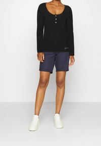 Zwarte geribbelde lange mouwen top met drie knopen, gecombineerd met marineblauwe shorts en witte sneakers. De shorts zijn omgeslagen aan de onderkant.