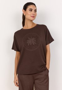 Braunes T-Shirt mit kurzen Ärmeln und rundem Halsausschnitt, mit einem kreisförmigen Palmbaum-Design und Text. Glatte Textur, lockere Passform.