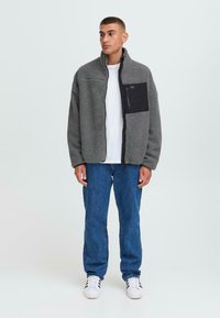 Gray fleece jacket with a black zippered pocket, high collar, and textured finish, paired with blue jeans and white sneakers.