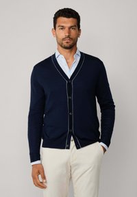 Uomo con un cardigan blu navy abbottonato sopra una camicia a righe azzurra chiara e pantaloni beige, in piedi contro uno sfondo chiaro e semplice.