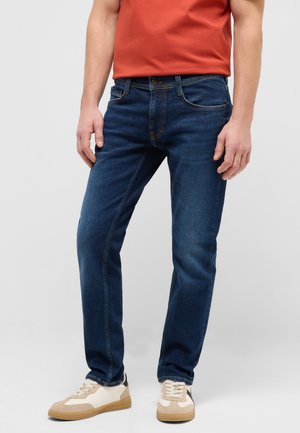 Mann trägt dunkelblaue Jeans, beige und weiße Sneakers und ein rotes Hemd, steht mit entspannten Händen an den Seiten.