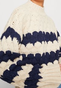 Paksu neulepaita, jossa on kerma- ja tummansinisiä sahalaitaisia raitoja, henkilöllä päällään. Paidassa on korkea ribbineck ja pitkät hihat.