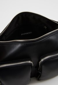 Borsa a mano in ecopelle nera con chiusura a zip, due tasche esterne con cerniere argentate e interno foderato. Etichetta del marchio visibile.