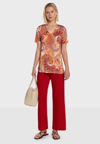Haut à manches courtes et col en V avec un imprimé cachemire vibrant aux tons orange, rouge et violet ; associé à un pantalon rouge à jambes larges et un sac à main clair.