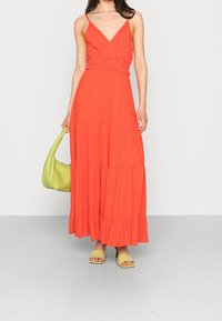 Robe maxi en orange vif avec un décolleté en V, un bustier ajusté et une jupe à étages. Associée à un sac à main vert clair et des sandales à talons larges.