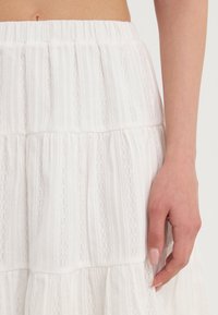 Gros plan sur la main d'une personne reposant à côté d'une jupe blanche texturée avec une taille élastique et des motifs de dentelle verticaux.