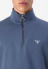 Μπλε ζακέτα με φερμουάρ και ψηλό γιακά, με ασημί φερμουάρ και μικρό κεντημένο λογότυπο στο στήθος. Μαλακό, υφασμένο ύφασμα.