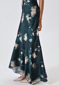 Abito in satin floreale con sfondo verde scuro, fiori crema chiaro e blu, orlo svasato, abbinato a scarpe beige con punta e cinturini alla caviglia.