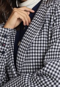 Žena v saku s černo-bílým vzorem houndstooth, pod kterým má modrou látku a bílý rolák, s rukou poblíž klíční kosti.