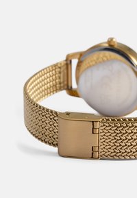 Χρυσό πλέγμα ρολόι χεριού με μεταλλικό λουρί, που διαθέτει λεία, γυαλιστερή επιφάνεια και κούμπωμα. Η πίσω πλευρά είναι καλυμμένη από το λουρί.