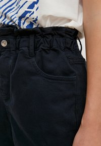 Pantalon en coton bleu marine foncé avec une taille froncée, des poches avant et un bouton-pression en métal. Texture lisse et coupe structurée.