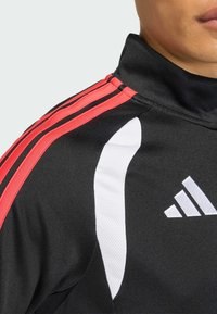 Μαύρο αθλητικό μπουφάν με κόκκινες και μαύρες ρίγες, λευκή λεπτομέρεια στον ώμο και λογότυπο Adidas. Το υλικό φαίνεται λείο και ελαφρώς υφασμένο.