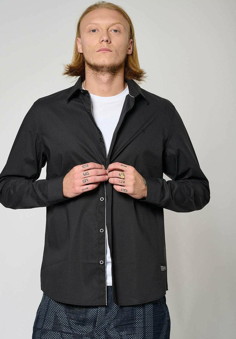 Zwarte lange mouwen shirt met knopen, met een witte binnenkraag en subtiele branding bij de zoom, gedragen over een wit t-shirt.