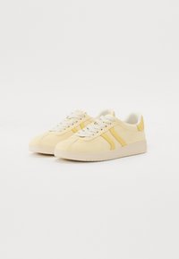 Gelbe und cremefarbene Sneakers mit einem glatten Wildleder-Obermaterial, weißen Schnürsenkeln, zwei gelben Streifen und einer strukturierten beigefarbenen Sohle.