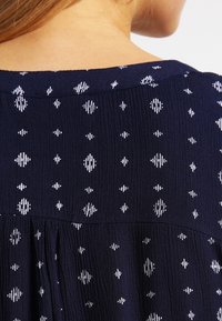 Mørkeblå stof med hvide diamant- og geometriske mønstre. Textureret overflade med lodrette linjer, rund halsudskæring og samlet detalje på ryggen.