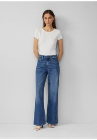 T-shirt blanc à côtes avec des manches à volants, associé à un jean bleu taille haute à jambes larges. Le mannequin porte des sandales à talons blanches.