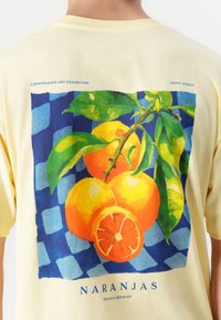T-shirt di cotone giallo con una stampa colorata di arance e foglie su uno sfondo a quadri blu, con un piccolo elemento di testo sottostante.