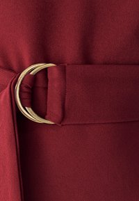 Vêtement ceinturé en tissu bordeaux, présentant une surface texturée. La quincaillerie en métal doré met en valeur le flux et la forme lisses du tissu.